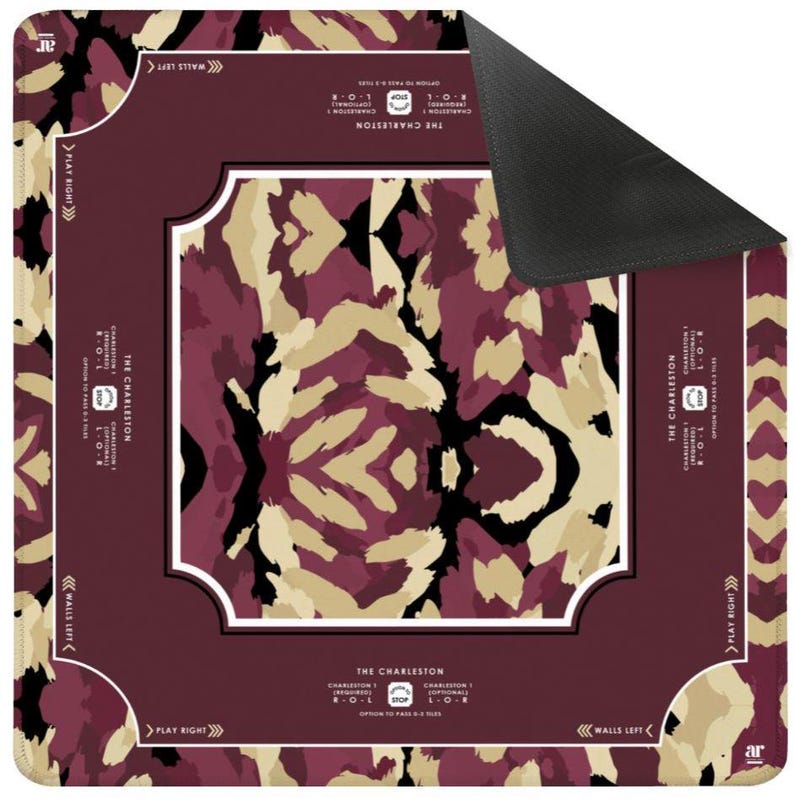 May include: A square game mat featuring a maroon, beige, and black abstract pattern. The border includes text reading THE CHARLESTON, CHARLESTON 1, R-O-L, OPTION TO PASS 0-3 TILES, WALLS LEFT, and PLAY RIGHT. One corner is folded back to reveal a black underside.