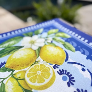 K&ouml;nnte beinhalten: Ein quadratisches dekoratives Tablett mit Zitronenmuster. Das Tablett hat einen blauen Rand mit einem wei&szlig;en und blauen Blumenmuster. Die Mitte des Tabletts zeigt gelbe Zitronen, wei&szlig;e Bl&uuml;ten und gr&uuml;ne Bl&auml;tter.