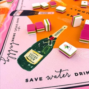 May include: A colorful Mahjong set with pink, white, and green tiles scattered on a pink and orange game board. The board features a champagne bottle illustration with the words "Lucky Pour" and "Save Water Drink Bubbly".