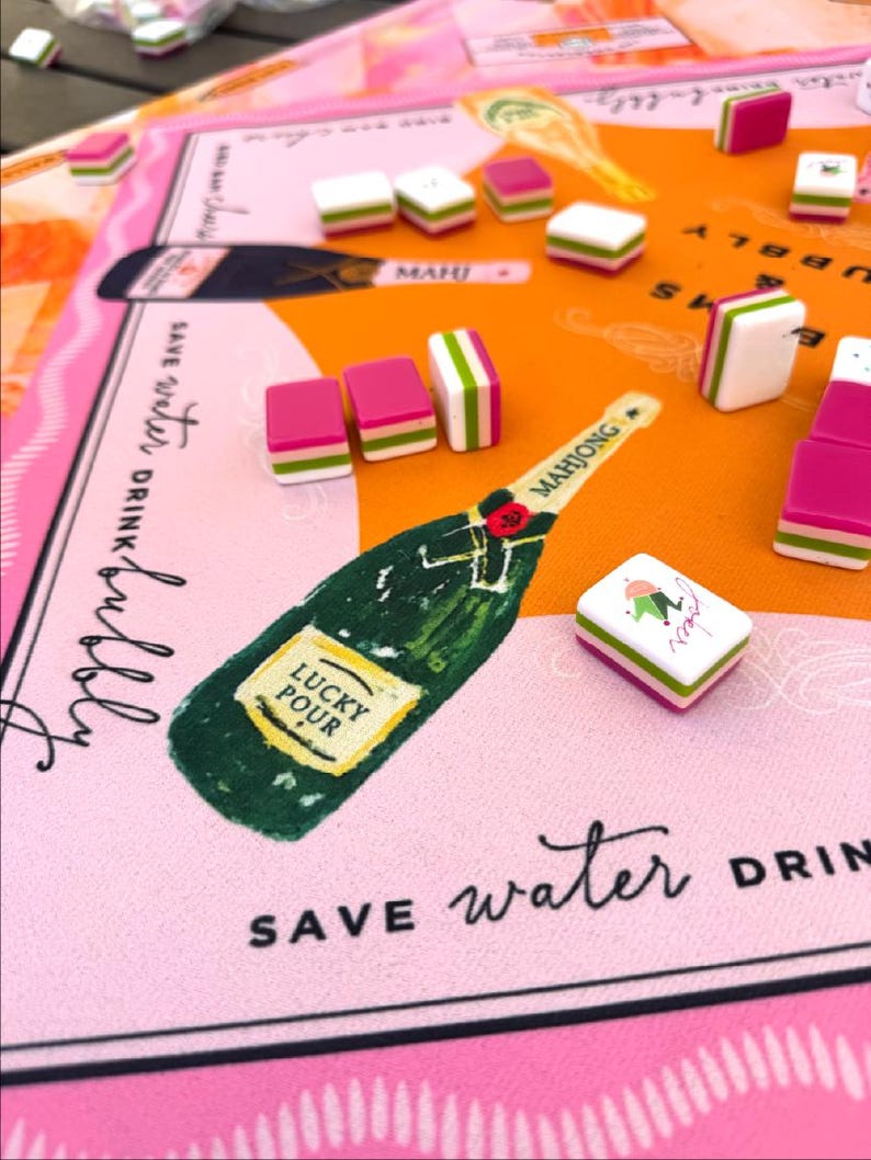 May include: A colorful Mahjong set with pink, white, and green tiles scattered on a pink and orange game board. The board features a champagne bottle illustration with the words "Lucky Pour" and "Save Water Drink Bubbly".