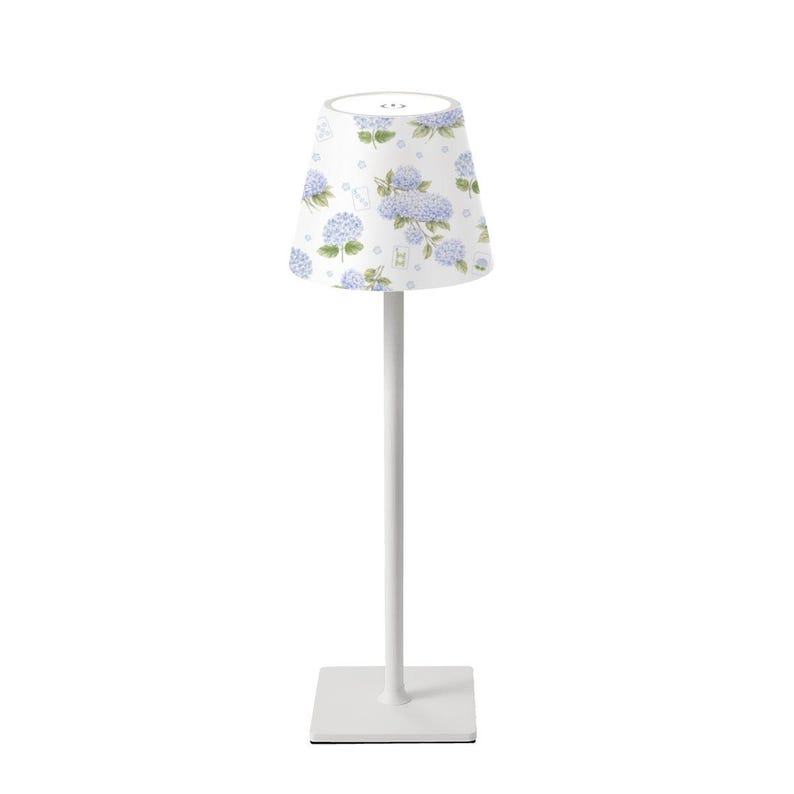K&ouml;nnte beinhalten: Eine wei&szlig;e Tischlampe mit quadratischem Fu&szlig; und schlankem Stiel. Der Lampenschirm ist wei&szlig; und mit einem Muster aus blauen und violetten Hortensienbl&uuml;ten verziert. Die Lampe ist f&uuml;r den Innenbereich konzipiert.
