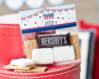 Op de afbeelding: Een rood, wit en blauwe cadeauzak met de tekst "happy 4th of JULY" gedrukt op de voorkant. De zak bevat een s'mores-set met graham crackers, marshmallows en een Hershey's chocolade reep.