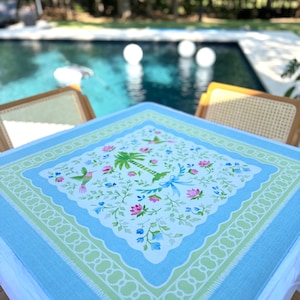May include: A square, light blue tablecloth with a floral and bird pattern. The center features a white background with pink, blue, and green flowers, palm trees, and hummingbirds. A green and white scalloped border frames the design.