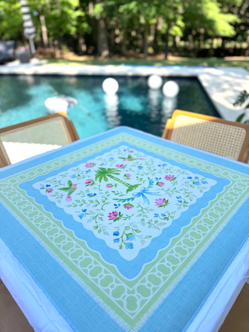 May include: A square, light blue tablecloth with a floral and bird pattern. The center features a white background with pink, blue, and green flowers, palm trees, and hummingbirds. A green and white scalloped border frames the design.