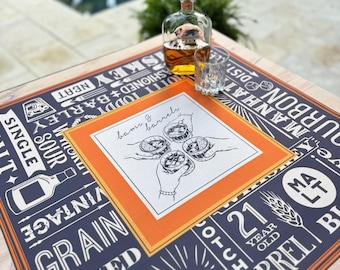 Op de afbeelding: Een tafelsetting met een whiskyfles en een glas. De tafel heeft een blauw en oranje patroonmat met tekst en illustraties over whisky. Het midden van de mat heeft een tekening van handen die whiskyglazen vasthouden. Een zwembad is op de achtergrond.