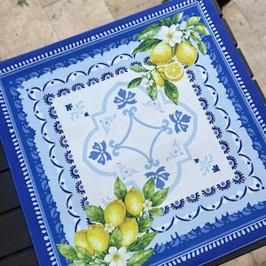 K&ouml;nnte beinhalten: Quadratische blaue und wei&szlig;e dekorative Tischdecke mit Zitronen- und Blumendesign. Der Rand ist dunkelblau mit einem gewellten Rand. Die Mitte zeigt ein hellblaues und wei&szlig;es Muster mit Zitronen und Blumen.