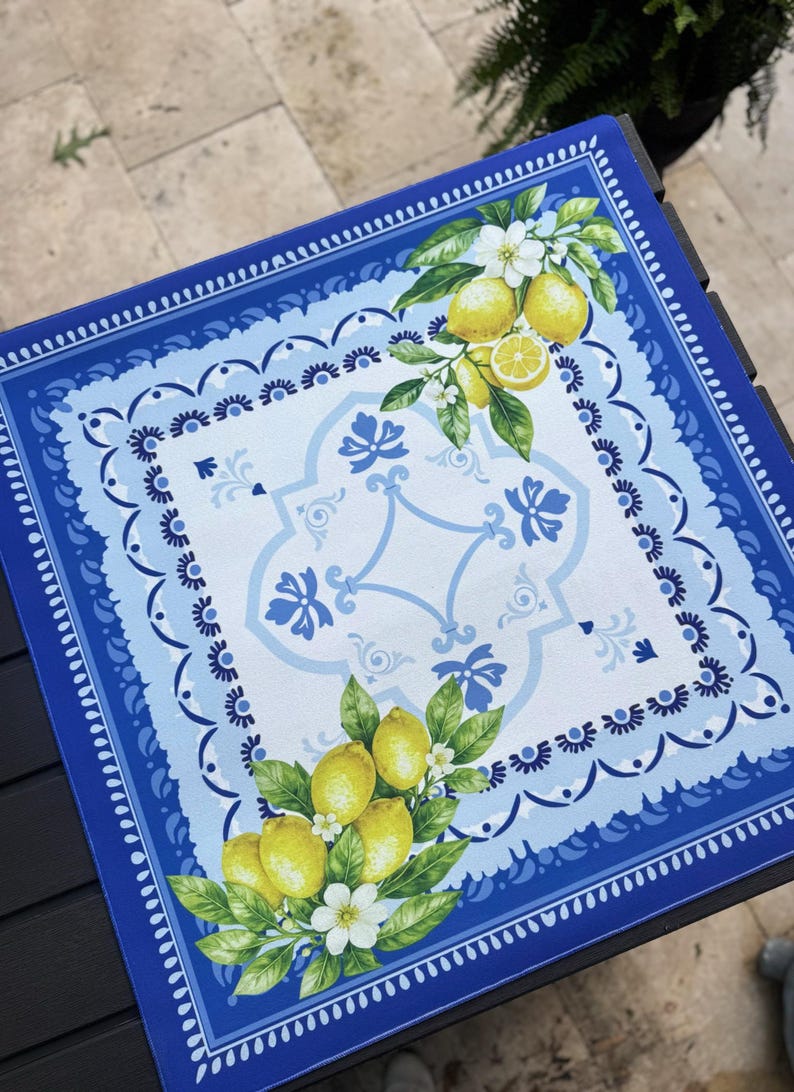 K&ouml;nnte beinhalten: Quadratische blaue und wei&szlig;e dekorative Tischdecke mit Zitronen- und Blumendesign. Der Rand ist dunkelblau mit einem gewellten Rand. Die Mitte zeigt ein hellblaues und wei&szlig;es Muster mit Zitronen und Blumen.