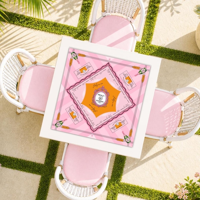 May include: Overhead view of a square white table set for outdoor dining, surrounded by four white wicker chairs with pink cushions. The table is covered with a pink and orange patterned tablecloth with champagne bottle illustrations.