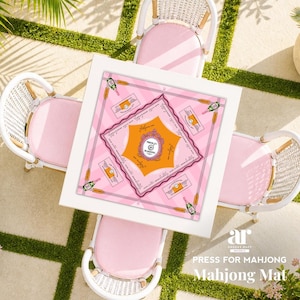 Pu&ograve; includere: Vista dall'alto di un tavolo quadrato bianco con un tappetino da Mahjong rosa e arancione. Il tappetino presenta un design a bottiglia di champagne e il testo "PRESS FOR MAHJONG". Quattro sedie in vimini bianche con cuscini rosa circondano il tavolo, su un patio.