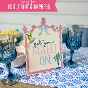 May include: A decorative sign with the text "get your MAHJ ON" in blue and pink, framed in pink, with a hummingbird and palm tree illustration. The sign is on a blue and white patterned tablecloth, with blue glasses in the foreground.
