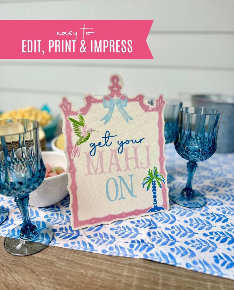 May include: A decorative sign with the text "get your MAHJ ON" in blue and pink, framed in pink, with a hummingbird and palm tree illustration. The sign is on a blue and white patterned tablecloth, with blue glasses in the foreground.