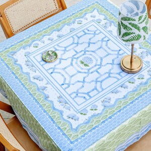 May include: A square tablecloth with a blue and green floral design, set on a table with wooden chairs. A small lamp with a gold base and a green fern pattern shade sits on the table. A small decorative plate is also on the table.