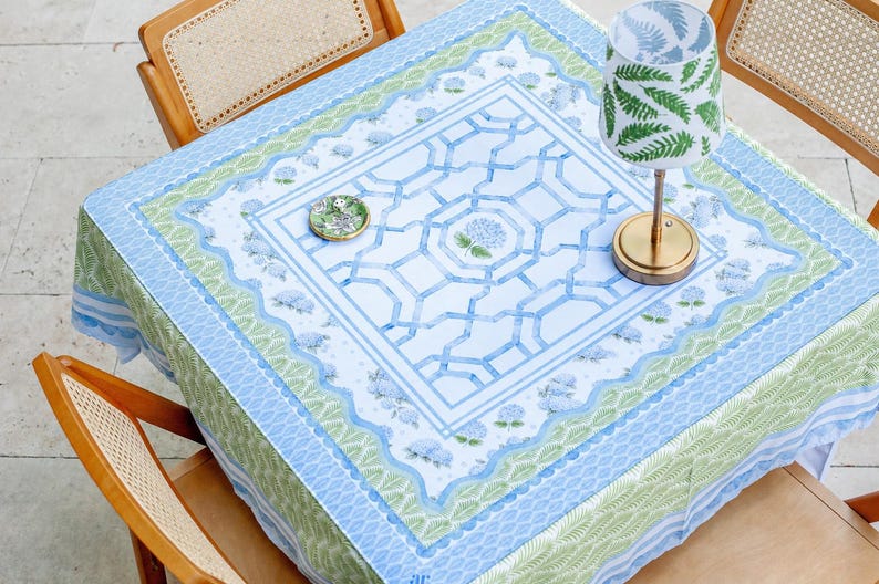 May include: A square tablecloth with a blue and green floral design, set on a table with wooden chairs. A small lamp with a gold base and a green fern pattern shade sits on the table. A small decorative plate is also on the table.
