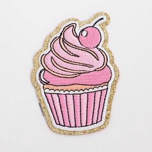 May include: Embroidered patch of a pink cupcake with a cherry on top. The cupcake has pink frosting with tan accents and a pink and white striped base. The patch is outlined with gold glitter.
