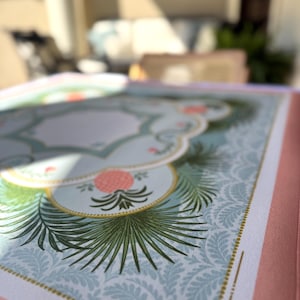 May include: Open book with a detailed illustration. The design features a light blue background with a repeating leaf pattern, accented by a decorative border and a central floral motif in pink and green. The book's edges are a soft pink.