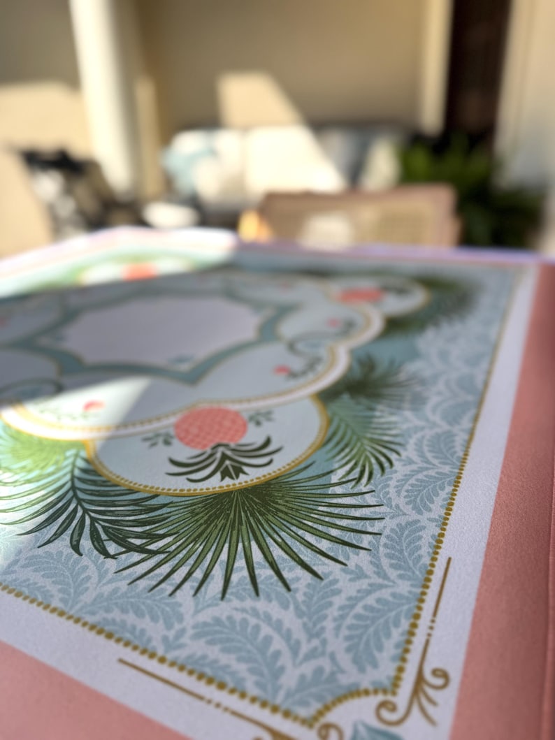 May include: Open book with a detailed illustration. The design features a light blue background with a repeating leaf pattern, accented by a decorative border and a central floral motif in pink and green. The book's edges are a soft pink.