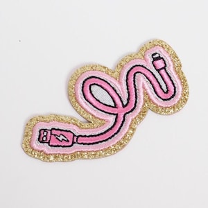 May include: Embroidered patch featuring a pink and black charging cable design with a lightning bolt symbol. The patch has a gold glitter border and is set against a white background. This patch is perfect for decorating clothing or accessories.