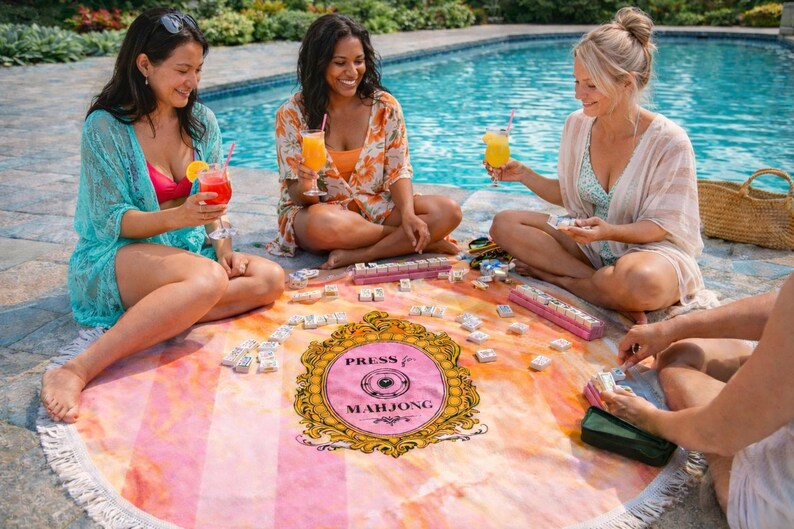 May include: A group of people are gathered around a round beach towel playing mahjong. The towel has a pink and orange watercolor design with the words "Press for Mahjong" in a gold frame. They are sitting near a pool.