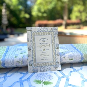 May include: A floral invitation for a "Pretty Little Mahjong Party" with text in a decorative frame. The invitation is light blue and white with hydrangea illustrations. The date is Friday, August 14th. The invitation is on a table with a blue and white floral pattern.