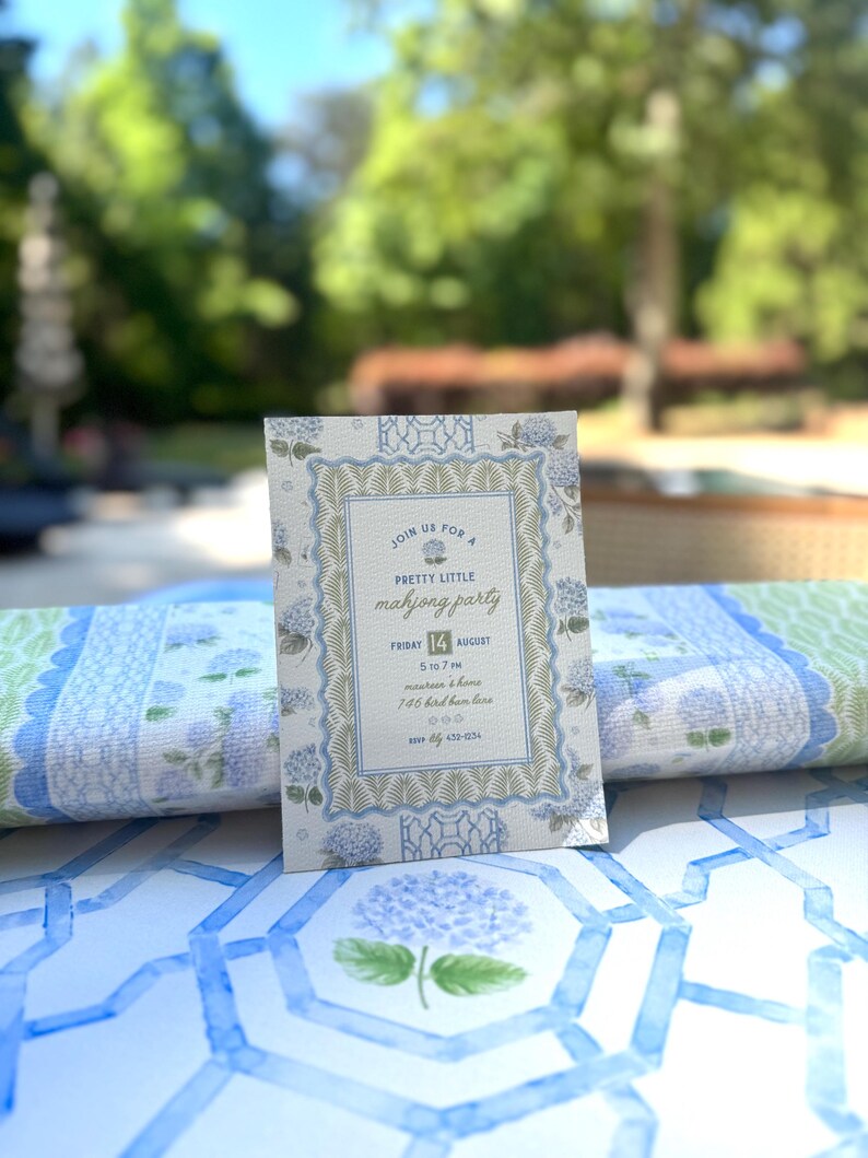May include: A floral invitation for a "Pretty Little Mahjong Party" with text in a decorative frame. The invitation is light blue and white with hydrangea illustrations. The date is Friday, August 14th. The invitation is on a table with a blue and white floral pattern.
