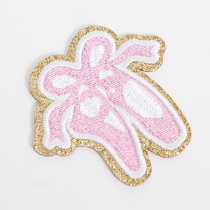 May include: Embroidered patch featuring a pair of pink ballet slippers with white accents. The patch has a gold glitter border and white stitching, perfect for adding to clothing or accessories.