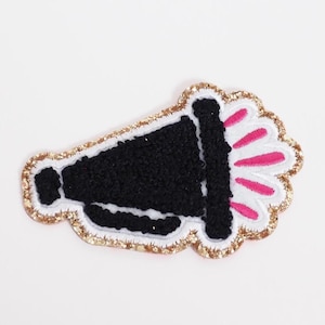 May include: A black megaphone patch with a white border and pink accents. The megaphone is outlined with gold glitter and has a textured appearance. This patch is suitable for jackets, bags, or other fabric items.