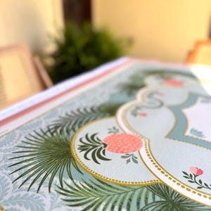 May include: Close-up of a decorative paper with a tropical design. The paper features a light blue background with a repeating pattern, accented with green palm leaves, pink flowers, and a gold-bordered central design.