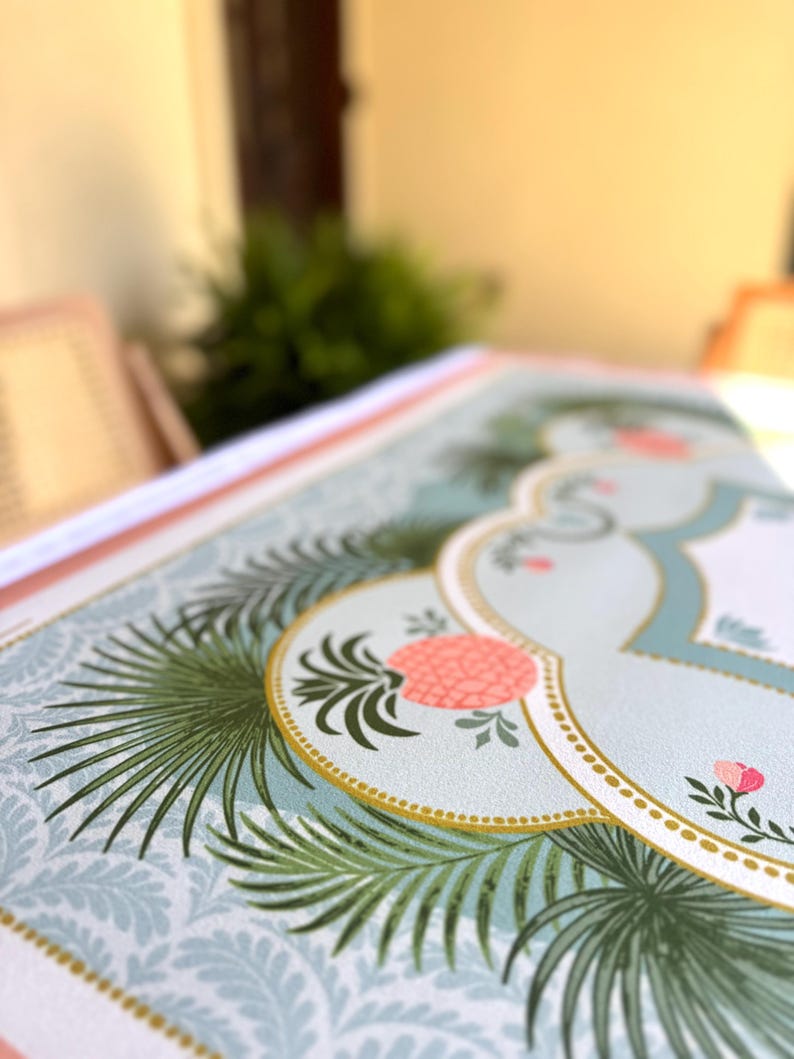 May include: Close-up of a decorative paper with a tropical design. The paper features a light blue background with a repeating pattern, accented with green palm leaves, pink flowers, and a gold-bordered central design.