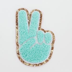 May include: A teal peace sign patch with a textured, fuzzy appearance. The patch is outlined with gold glitter and has a white border. The peace sign is a symbol of unity and harmony.