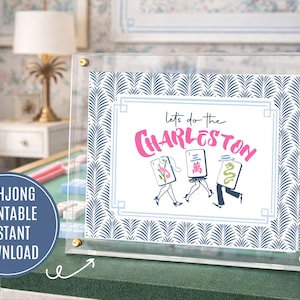 Puede incluir: Una impresi&oacute;n enmarcada con el texto "Let's do the Charleston" en rosa, con figuras de dibujos animados con fichas de Mahjong. La impresi&oacute;n tiene un patr&oacute;n de hojas azul y blanco y se muestra sobre una superficie verde.