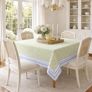 K&ouml;nnte beinhalten: Ein Esstisch mit einer hellgr&uuml;nen Tischdecke mit wei&szlig;em und blauem Rand. Vier wei&szlig;e St&uuml;hle mit gewebten R&uuml;cken umgeben den Tisch. Ein Blumenarrangement und Kerzen stehen auf dem Tisch.