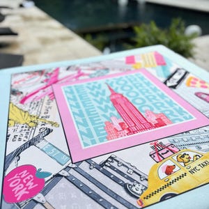 May include: A light blue square print featuring a pink framed image of the Empire State Building with the text "NEW YORK" and "NYC TAXI". The print also includes illustrations of luggage, a yellow taxi, and street signs.