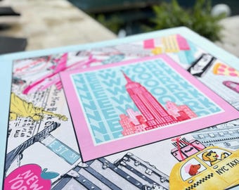 Puede incluir: Una impresión cuadrada azul claro con una imagen enmarcada en rosa del Empire State Building, el texto "NEW YORK" y "NYC TAXI". La impresión también incluye ilustraciones de equipaje, un taxi amarillo y señales de tráfico.