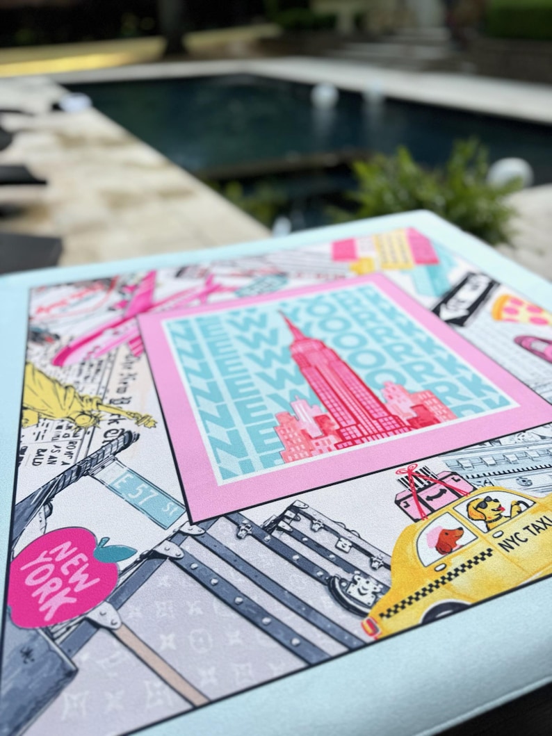 May include: A light blue square print featuring a pink framed image of the Empire State Building with the text "NEW YORK" and "NYC TAXI". The print also includes illustrations of luggage, a yellow taxi, and street signs.
