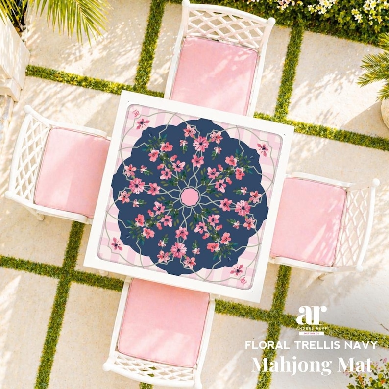 May include: Overhead view of a white square table with a navy blue floral design and four white chairs with pink cushions. The table has the text "FLORAL TRELLIS NAVY Mahjong Mat". The chairs are arranged around the table on a stone patio.
