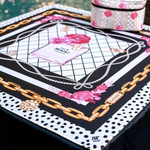 May include: A square black and white scarf with a gold chain design and pink floral accents. The scarf features a perfume bottle illustration with the text "EAU DE MAHU". A small, matching cosmetic bag sits on top.