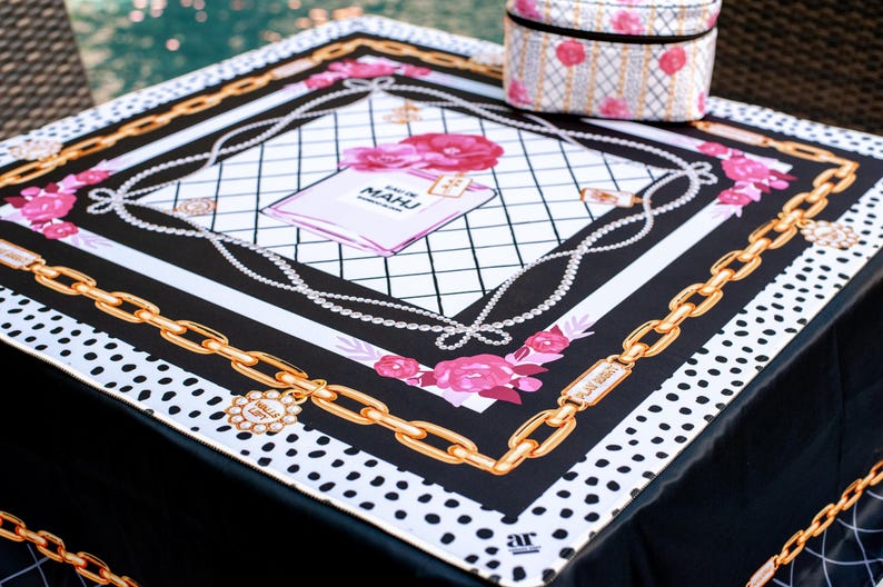 May include: A square black and white scarf with a gold chain design and pink floral accents. The scarf features a perfume bottle illustration with the text "EAU DE MAHU". A small, matching cosmetic bag sits on top.