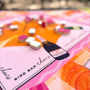 May include: A close-up of a board game with a pink and orange color scheme. The game board features the text "BIRD BAM Cheers" and a champagne bottle illustration. Small, colorful game pieces are scattered across the board.