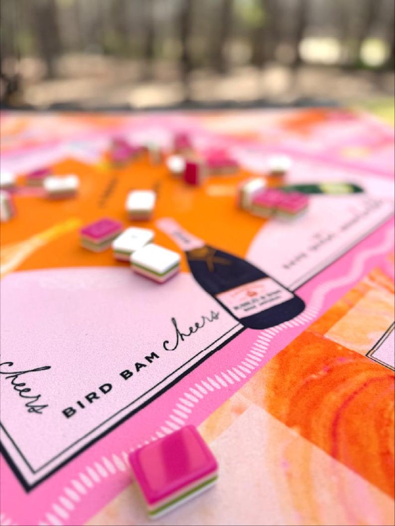 May include: A close-up of a board game with a pink and orange color scheme. The game board features the text "BIRD BAM Cheers" and a champagne bottle illustration. Small, colorful game pieces are scattered across the board.
