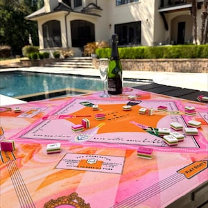 May include: A pink and orange Mahjong game set on a table with a bottle of champagne and a glass. The game board has text that reads "Press for Mahjong" and "Save water, drink bubbly."