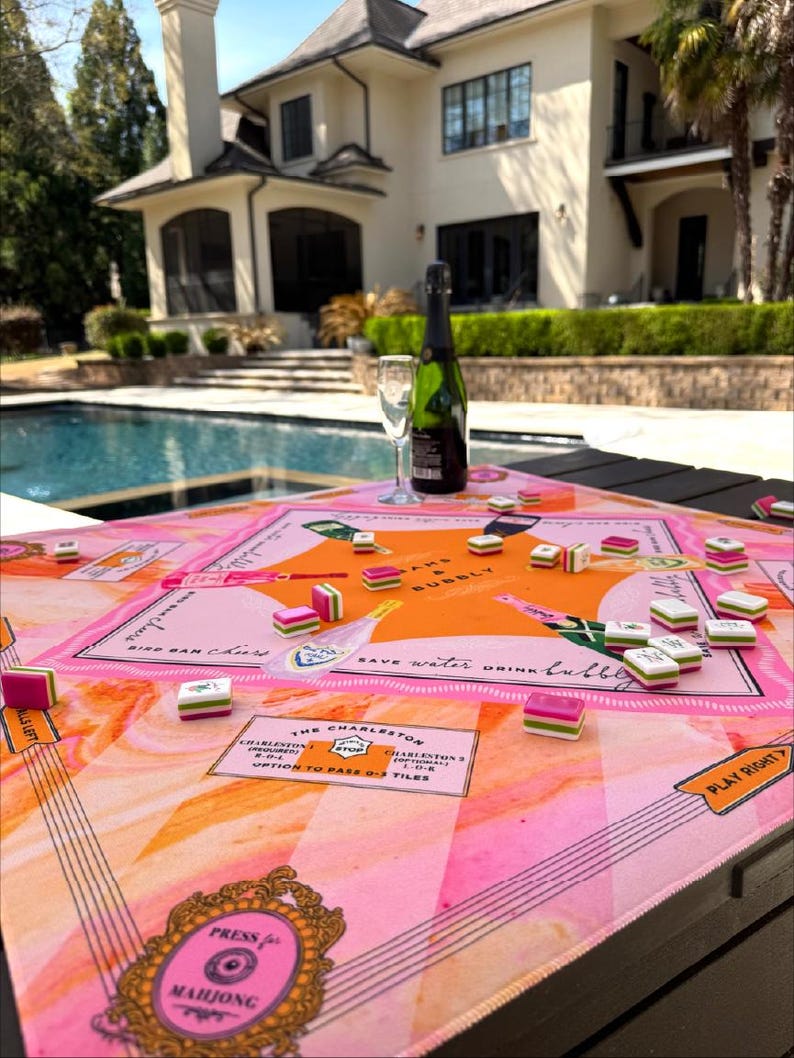 May include: A pink and orange Mahjong game set on a table with a bottle of champagne and a glass. The game board has text that reads "Press for Mahjong" and "Save water, drink bubbly."
