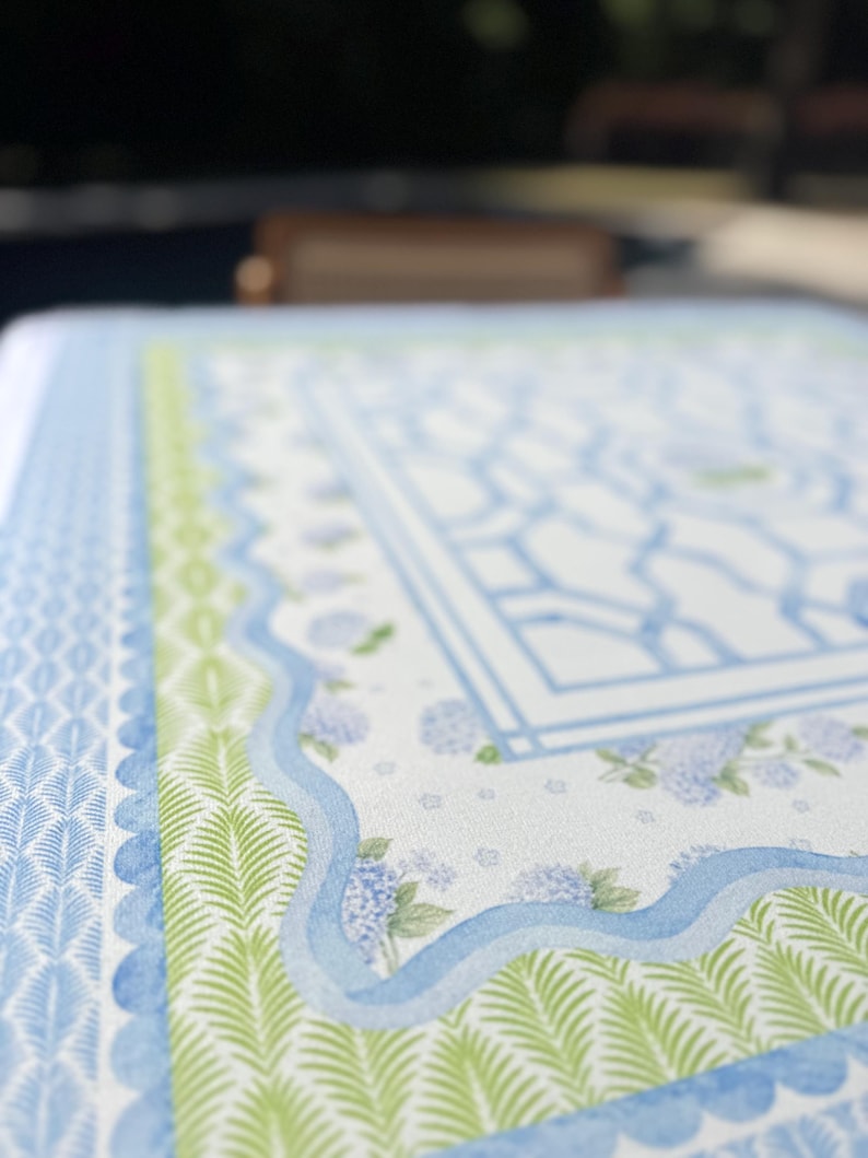 May include: Close-up of a patterned tablecloth with a blue and green design. The tablecloth features a repeating pattern of blue and white geometric shapes, green fern-like leaves, and blue scalloped edges. Hydrangea flowers are also visible.