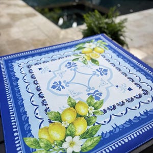 K&ouml;nnte beinhalten: Quadratisches blau-wei&szlig;es Dekotablett mit Zitronen- und Blumenmuster. Das Tablett hat einen gewellten Rand und ein zentrales Muster mit Zitronen, Bl&auml;ttern und wei&szlig;en Bl&uuml;ten. Im Hintergrund ein Swimmingpool.