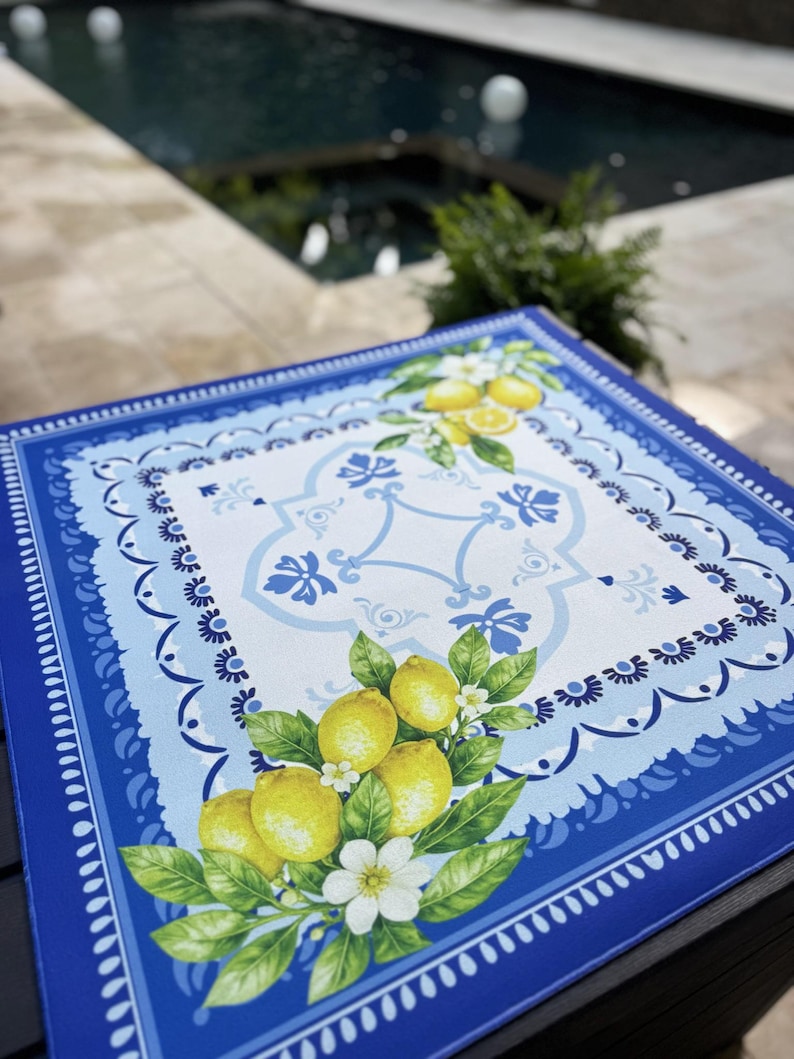 K&ouml;nnte beinhalten: Quadratisches blau-wei&szlig;es Dekotablett mit Zitronen- und Blumenmuster. Das Tablett hat einen gewellten Rand und ein zentrales Muster mit Zitronen, Bl&auml;ttern und wei&szlig;en Bl&uuml;ten. Im Hintergrund ein Swimmingpool.
