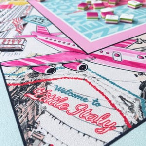 May include: A board game with a travel theme, featuring a pink airplane, the words "Welcome to Little Italy," and game pieces. The game board has a light blue background and a pink border. The game pieces are pink, white, and green.