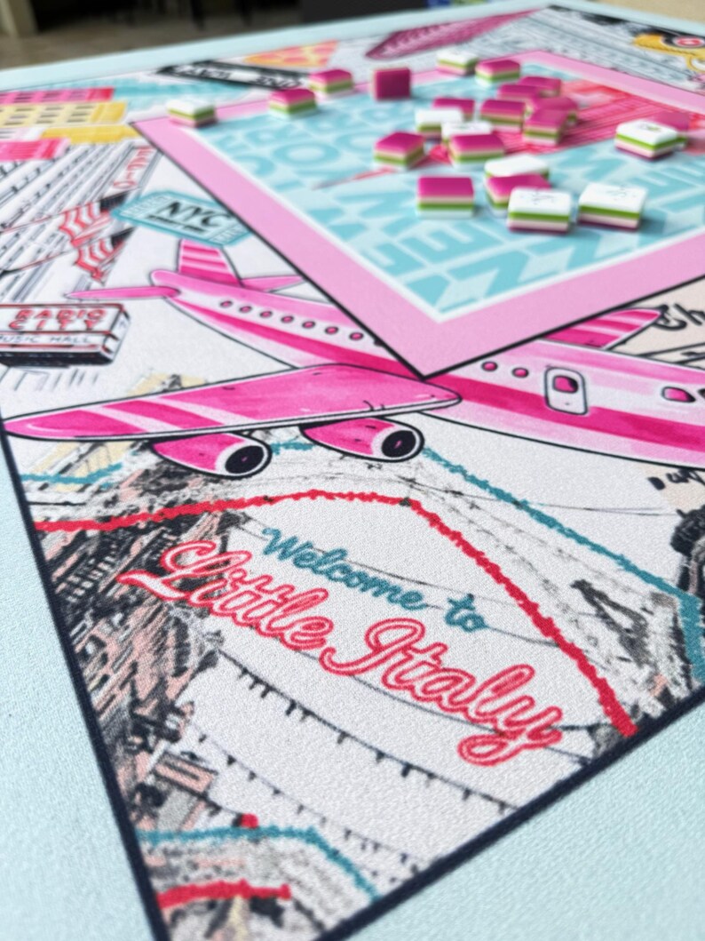 May include: A board game with a travel theme, featuring a pink airplane, the words "Welcome to Little Italy," and game pieces. The game board has a light blue background and a pink border. The game pieces are pink, white, and green.