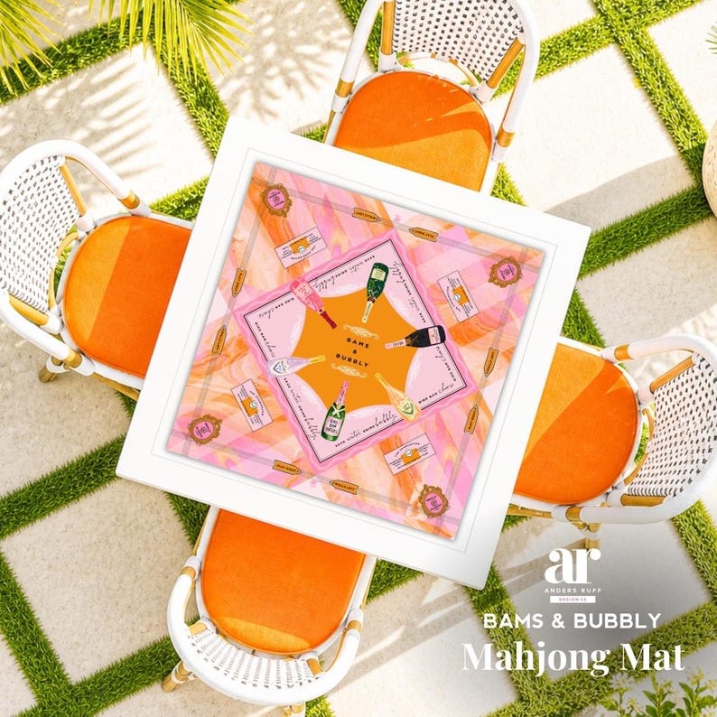 May include: Overhead shot of a white square table with a pink and orange Mahjong mat, surrounded by four white wicker chairs with orange cushions. The mat features champagne bottles and the text "BAMS & BUBBLY".