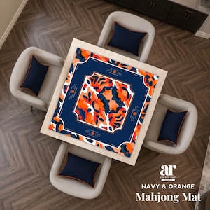 May include: A square mahjong mat featuring a central abstract pattern of orange, white, and dark blue shapes framed by a solid dark blue border with white text accents. The mat is centered on a light wood table surrounded by four chairs with dark blue cushions.