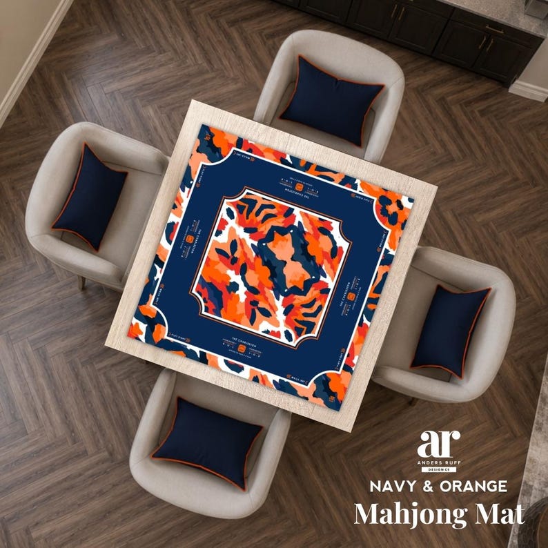 May include: A square mahjong mat featuring a central abstract pattern of orange, white, and dark blue shapes framed by a solid dark blue border with white text accents. The mat is centered on a light wood table surrounded by four chairs with dark blue cushions.