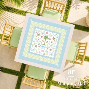 May include: A square Mahjong mat with a light blue border and a central design featuring a palm tree, hummingbirds, and floral elements. The mat is set on a table with bamboo chairs and green cushions.